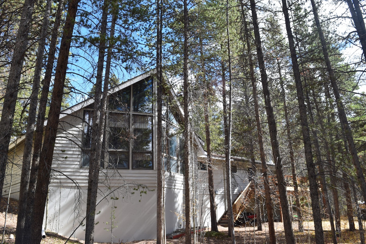 The exterior of the mountain home is surrounded by tall pine trees, showcasing large windows that offer plenty of natural light. The structure features a modern design, with a mix of wood and siding, blending harmoniously with the natural landscape.