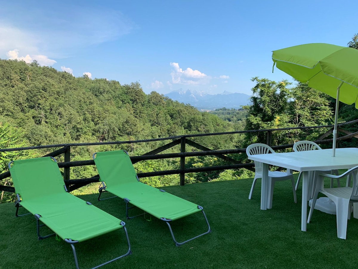 Two green lounge chairs are positioned on a grassy area, complemented by a white table and chairs set under a large umbrella. A scenic view of rolling hills and distant mountains is visible in the background, framed by lush greenery.