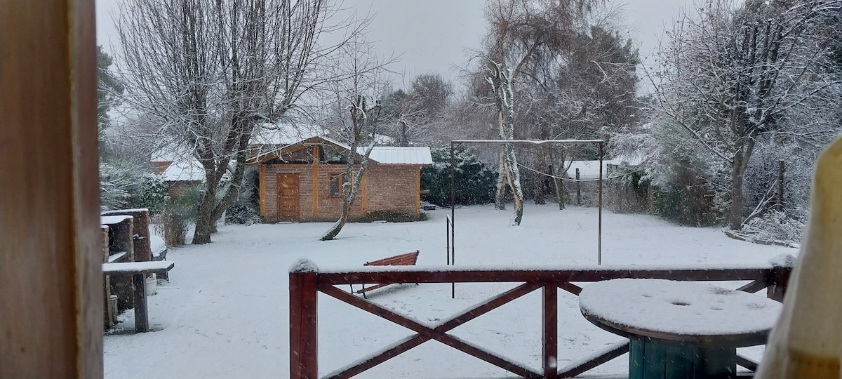 A snowy landscape is visible from a wooden porch, featuring a charming cabin surrounded by trees. The yard is blanketed in snow, while a picnic table and a clothesline stand in the foreground, providing a serene wintry scene.