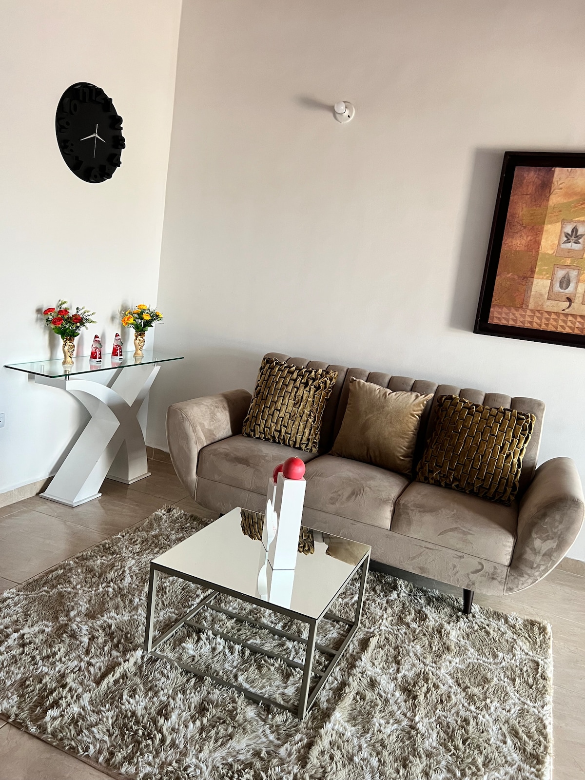 A cozy living area is presented with a plush sofa adorned with decorative cushions, accompanied by a glass coffee table. A side table displays small floral arrangements, and a large artwork adds visual interest to the wall. Natural light enhances the serene atmosphere.
