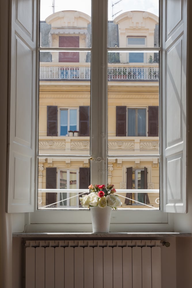 A bright window opens to a view of a charming building with a decorative facade, showcasing balconies and shuttered windows. A vase with flowers rests on the windowsill, adding a touch of nature to the indoor setting.