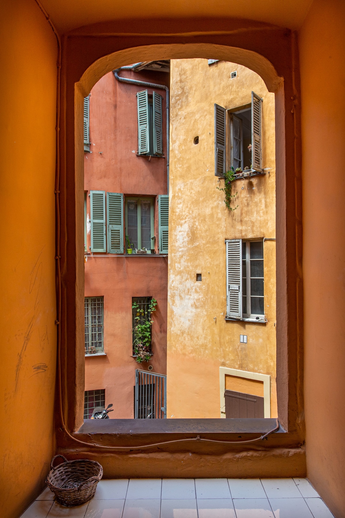 A view from a doorway showcases vibrant, contrasting colors of adjoining buildings. The orange and yellow facades are complemented by green shutters and window boxes with plants. A small woven basket rests on the tiled floor, enhancing the scene's character.