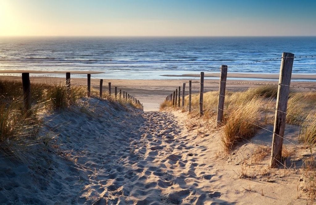 A sandy path leads to the beach, framed by grass and wooden posts. The tranquil sea horizon meets the sky at sunset, providing a serene view of the ocean waves.