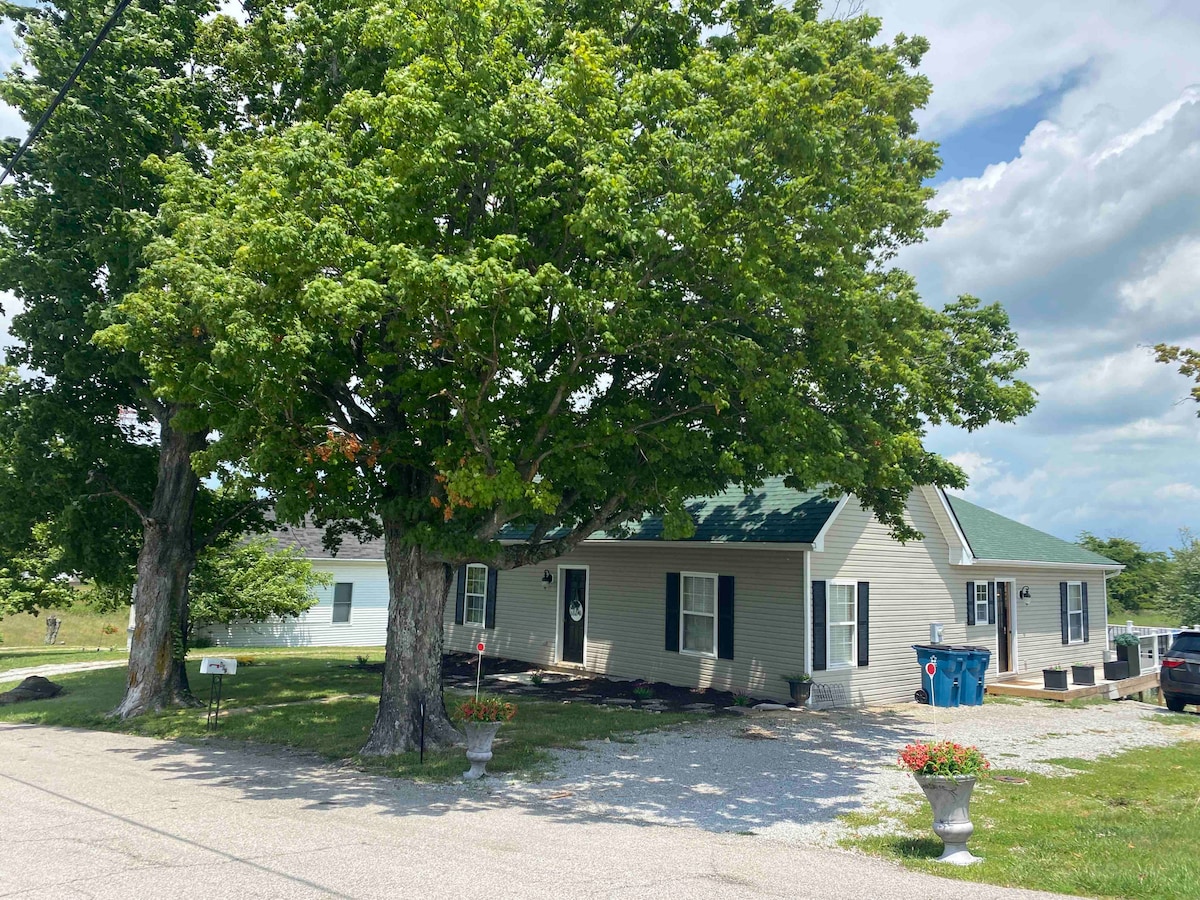 A spacious house surrounded by mature trees and well-maintained landscaping is depicted. A gravel driveway leads to the entrance, and colorful flower pots add a welcoming touch. Clear skies and distant hills can be seen in the background, enhancing the serene setting.
