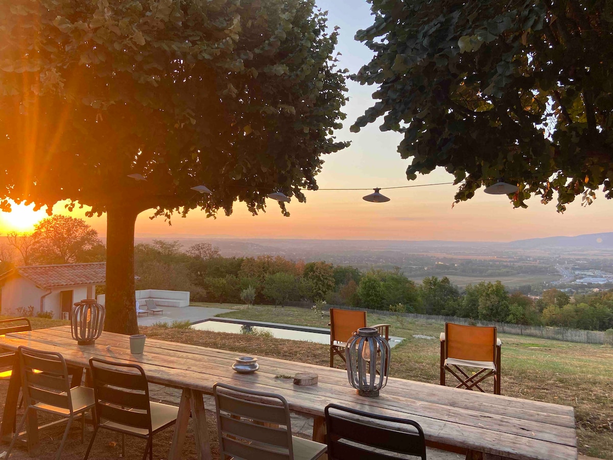 A spacious outdoor dining area is set beneath large trees, featuring a long wooden table surrounded by chairs. Soft light from the sunset casts a warm glow across the landscape, which includes a swimming pool and distant hills, creating a serene atmosphere.