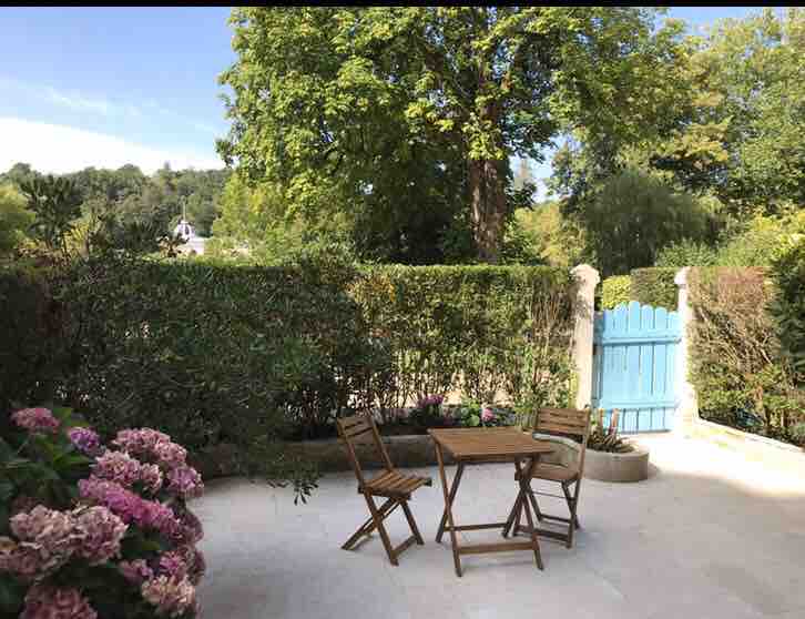 An outdoor area features a small wooden table with two chairs, surrounded by lush greenery. A vibrant blue garden gate can be seen in the background, complemented by blooming flowers in shades of pink nearby. The space is bright and sunny, enhancing its inviting ambiance.