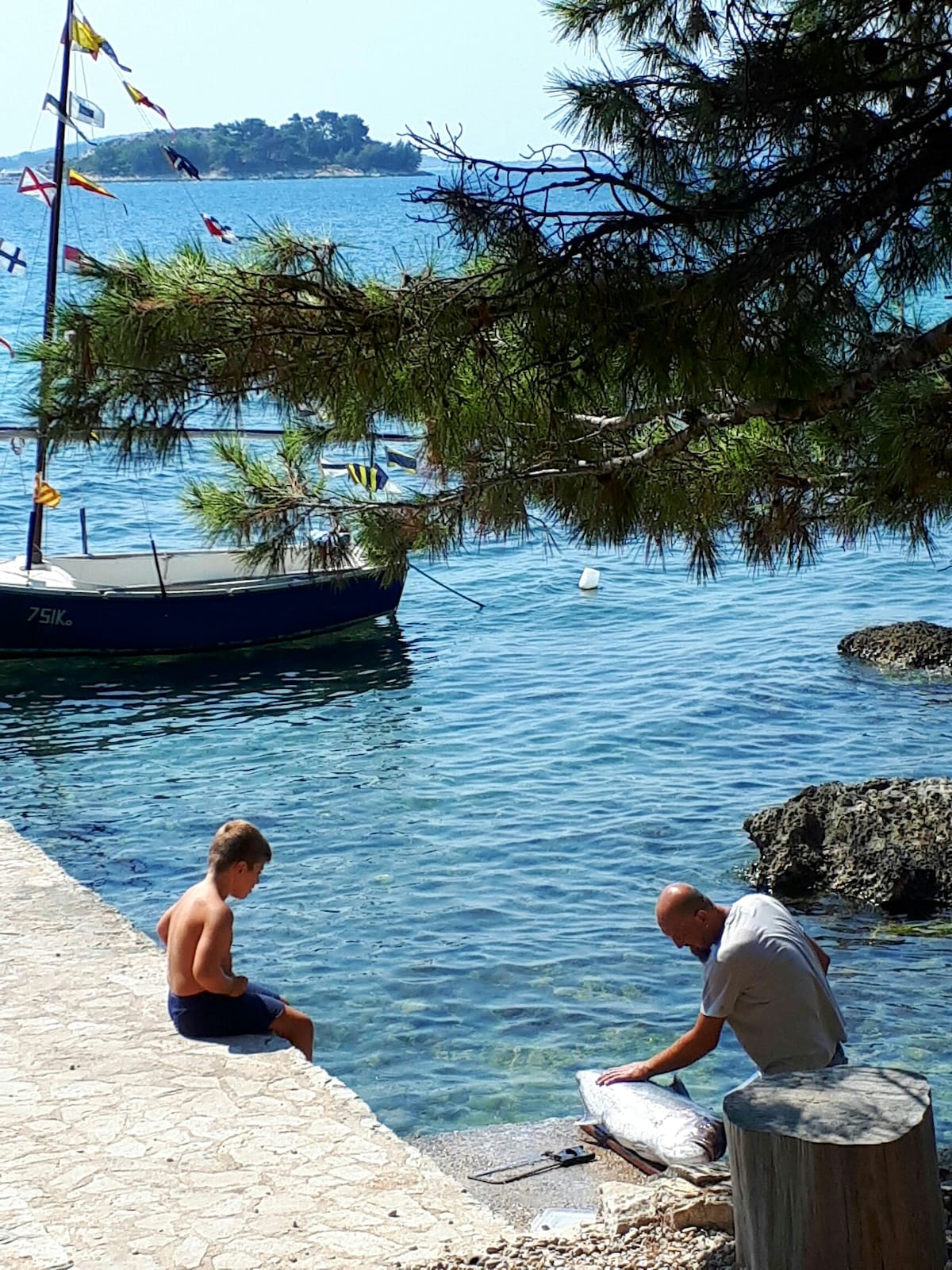 A calm seaside scene is captured, featuring a child seated on a stone pathway, observing an adult beside a fishing boat. Clear blue water reflects nearby greenery and boats are anchored in the distance, creating a serene coastal atmosphere.