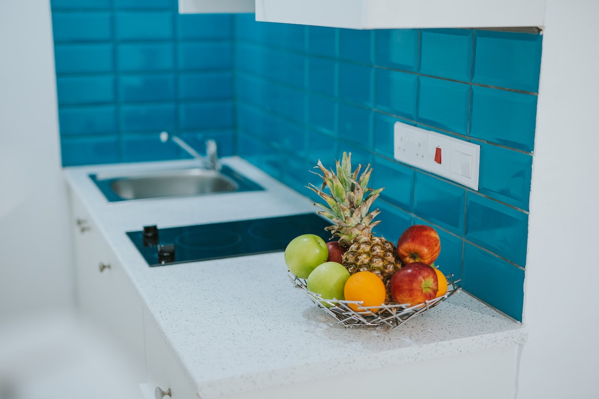 A kitchenette area displays a compact cooking space, featuring a sink and a stovetop. A vibrant blue tiled backsplash complements the light countertop. A bowl of assorted fresh fruits, including a pineapple, apples, and oranges, is positioned prominently, enhancing the space's functionality.