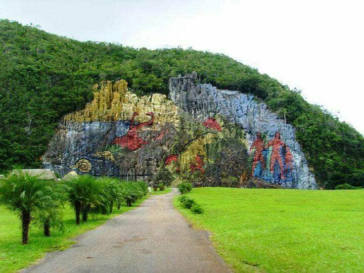 A colorful mural is painted on a rocky cliffside, depicting various figures and symbols. Lush greenery is present in the foreground, accompanied by short tree-like plants. A pathway leads towards the mural, inviting exploration of the natural surroundings and the art.