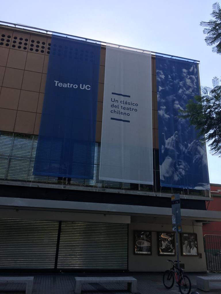 The exterior of Teatro UC is shown, featuring large blue banners with the words 'Teatro UC' and 'Un clásico del teatro chileno'. The building has a modern design, with glass windows at the ground level and a tree partially visible to the side.