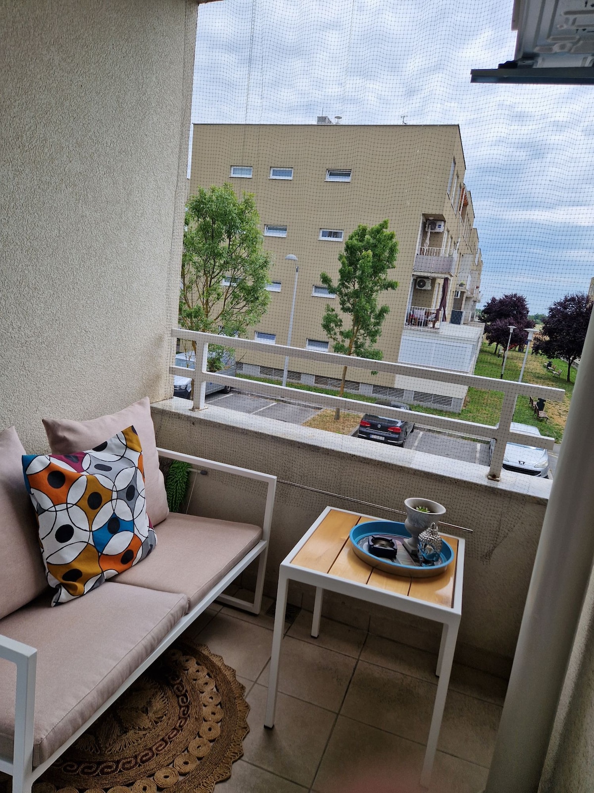 A cozy terrace area features a light-colored sofa adorned with a patterned cushion. A small wooden table holds decorative items. The view includes greenery and parked cars, framed by the building structure and soft sky tones.