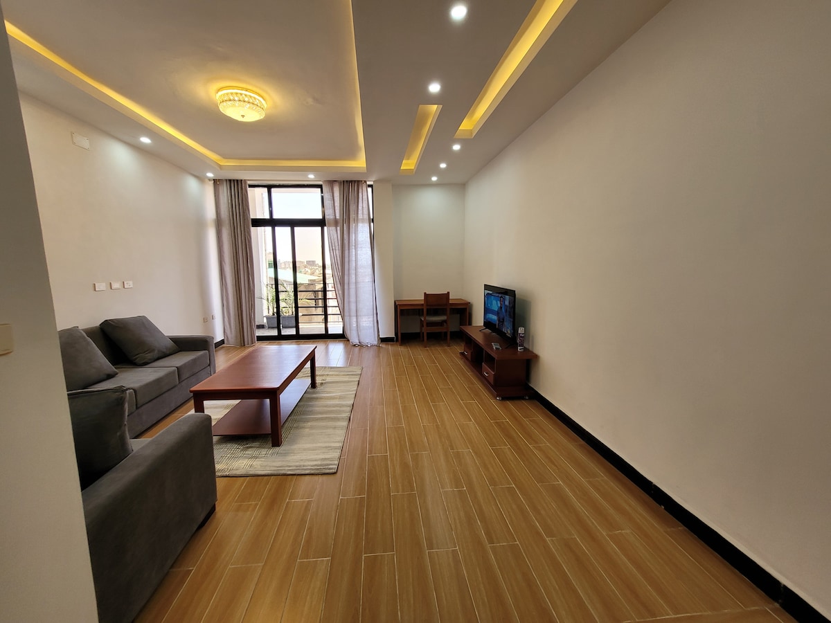 A modern living area is presented with a comfortable gray sofa and a wooden coffee table. Large sliding glass doors provide natural light and access to a balcony. A smart TV is positioned on a media console against the wall, complemented by a workspace with a chair.