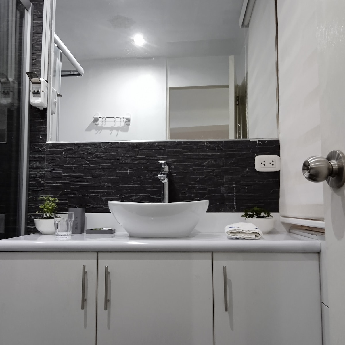 A modern bathroom is presented with a clean white sink positioned on a light countertop. Black stone tiles create a textured backdrop. Soft green plants are placed on either side of the sink, and a towel is neatly rolled beside a small glass. Natural light is visible.