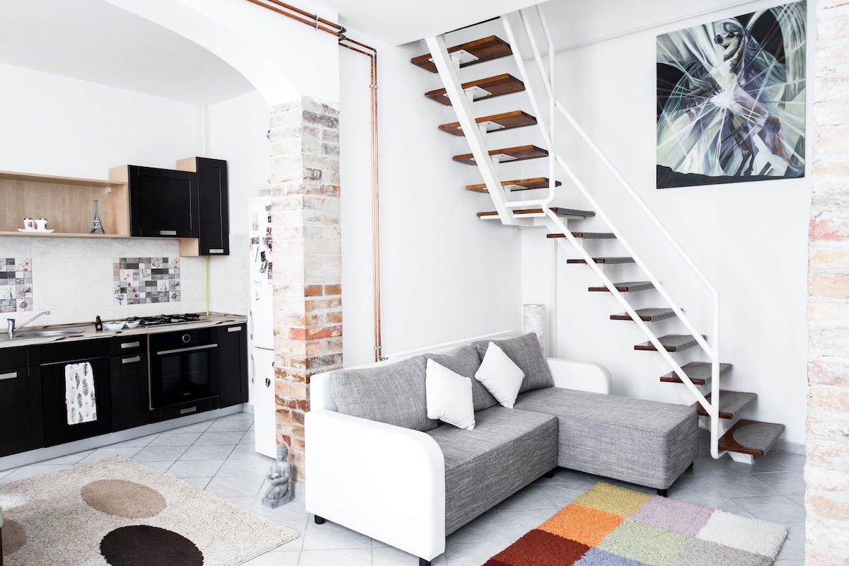 A bright living area features a modern gray sofa with white accents and four decorative pillows. A colorful area rug is positioned beneath it. The kitchen area is visible, showcasing dark cabinetry and a tiled backsplash. A stylish staircase leads to the upper level.