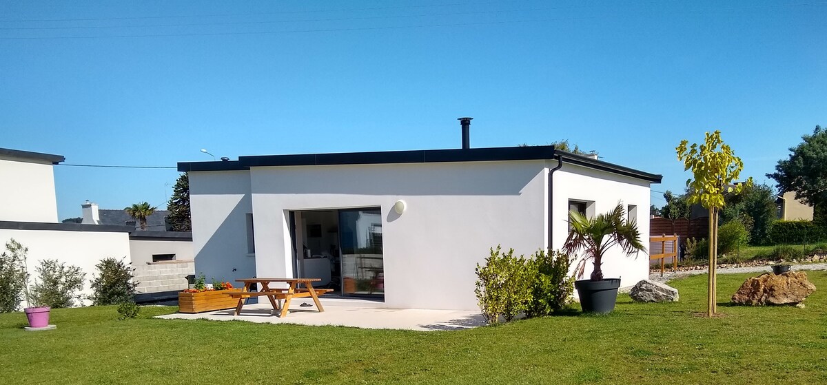 A contemporary house exterior is displayed with a smooth white facade. Large glass doors open to a terrace area, complemented by a picnic table and greenery. The surrounding lawn is well-kept with various plants and a clear blue sky visible above.