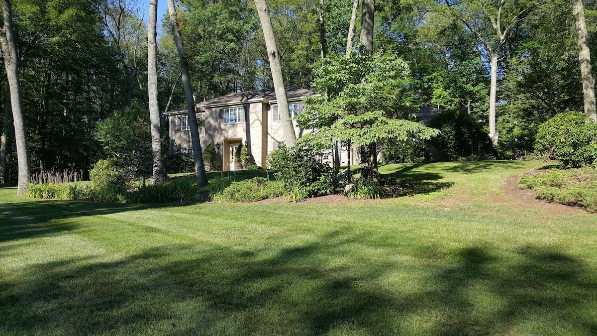 The exterior of a two-story home is visible, surrounded by lush greenery and trees. A manicured lawn leads to the house, showcasing a blend of natural landscaping and structured plantings. The facade features large windows, allowing for an abundance of natural light.