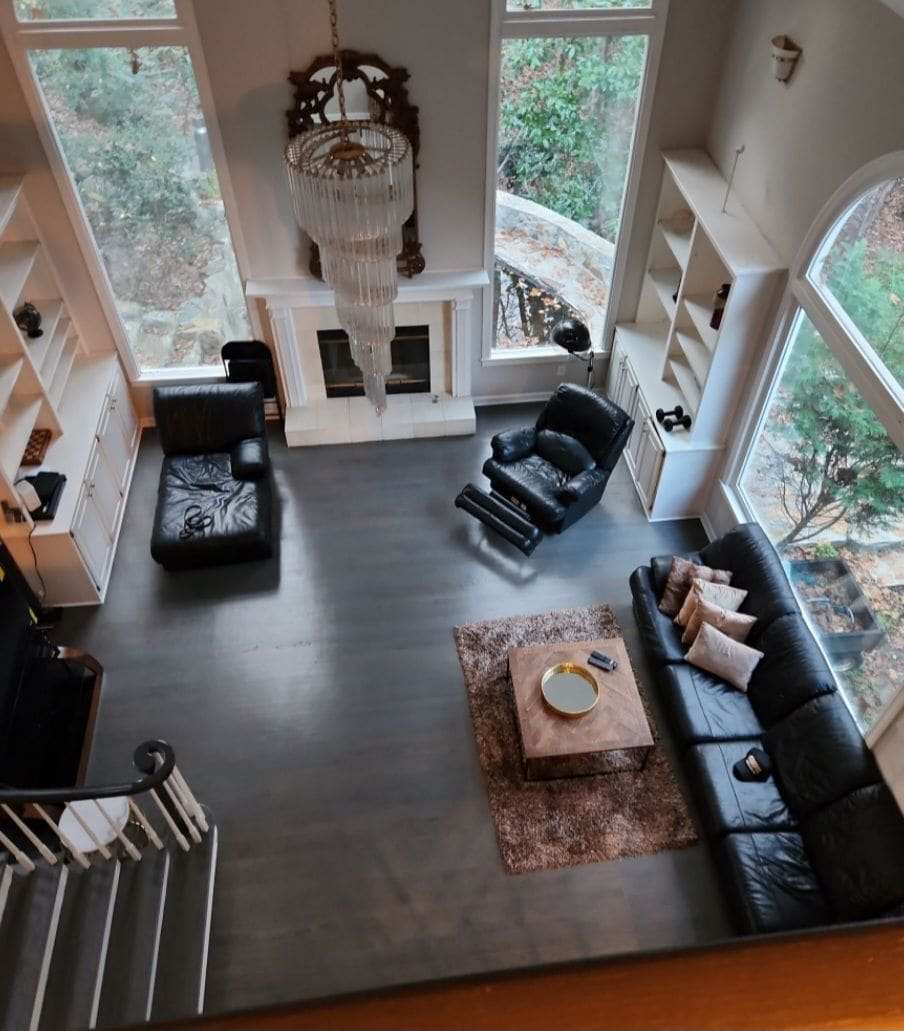A spacious living area is featured, with large windows allowing natural light to fill the room. Comfortable black leather seating is arranged around a circular coffee table on a soft area rug. A chandelier hangs above, complementing the fireplace and built-in bookshelves.