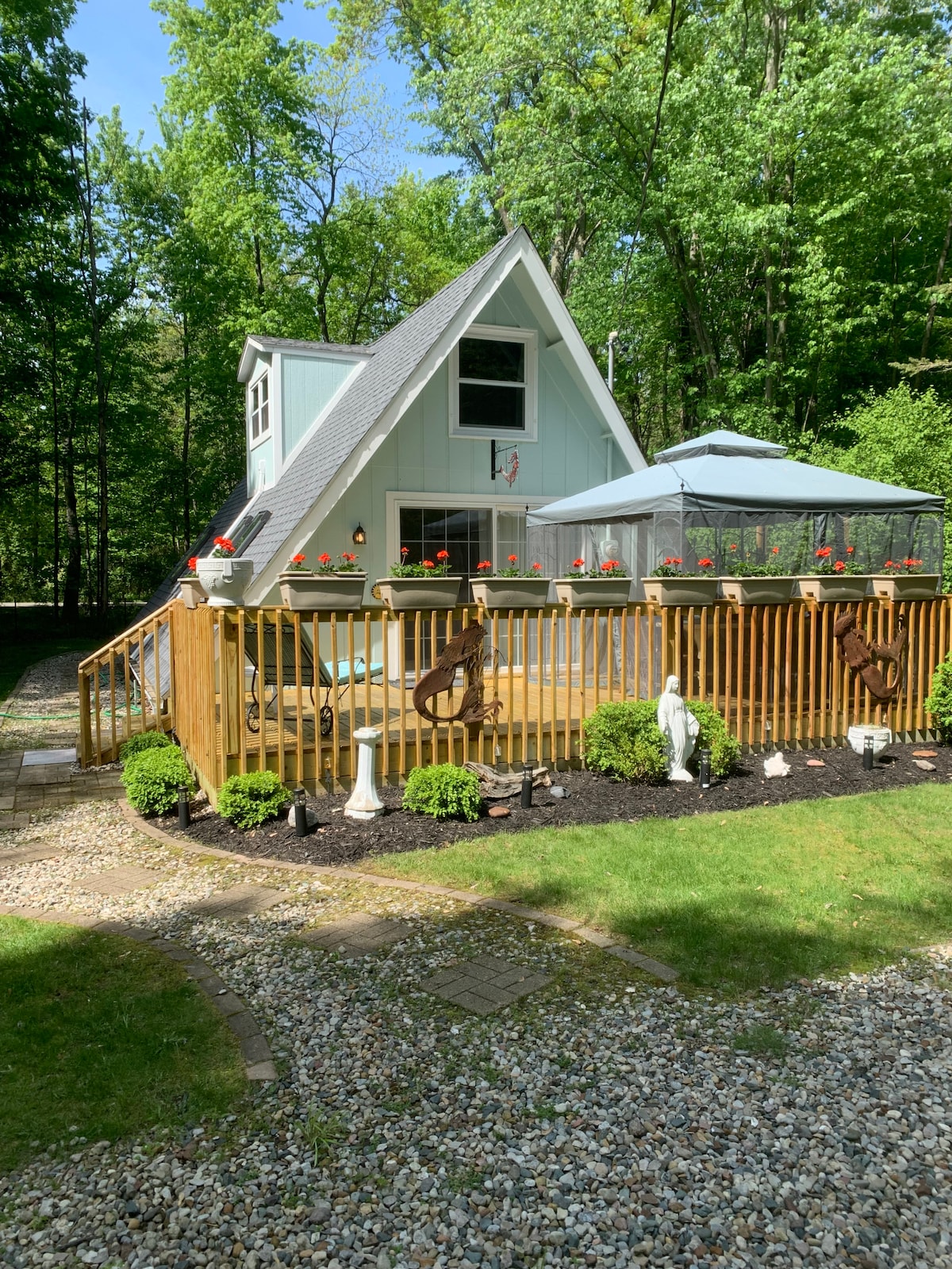 A charming cottage is surrounded by lush greenery, featuring an inviting deck enclosed by a wooden fence. Potted flowers add color, while decorative elements and a gazebo enhance the outdoor space. A stone pathway leads to the entrance, creating a welcoming ambiance.