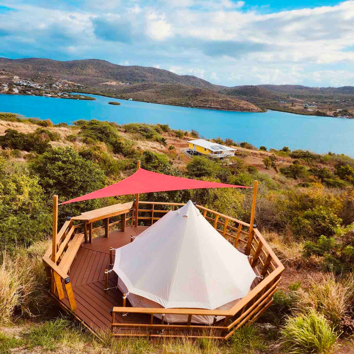 A spacious wooden deck surrounds a large, white tent set against a backdrop of lush greenery and tranquil blue waters. The serene landscape features rolling hills and distant houses, with a vibrant canopy providing shade. The scene captures a sense of calm and natural beauty.