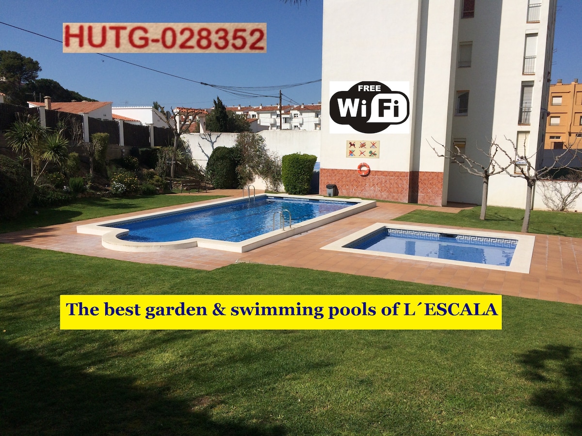 A well-maintained garden area features two swimming pools surrounded by manicured grass. One pool is designed for children, while the larger pool includes a Roman staircase for easy access. Clear blue water reflects sunlight, creating a refreshing setting for relaxation.