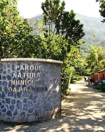 A stone entrance sign marks the Parque Natural Municipal da Prainha, surrounded by lush greenery. A pathway leads into the park, with a gentle slope visible in the background, suggesting a natural setting ideal for exploration.