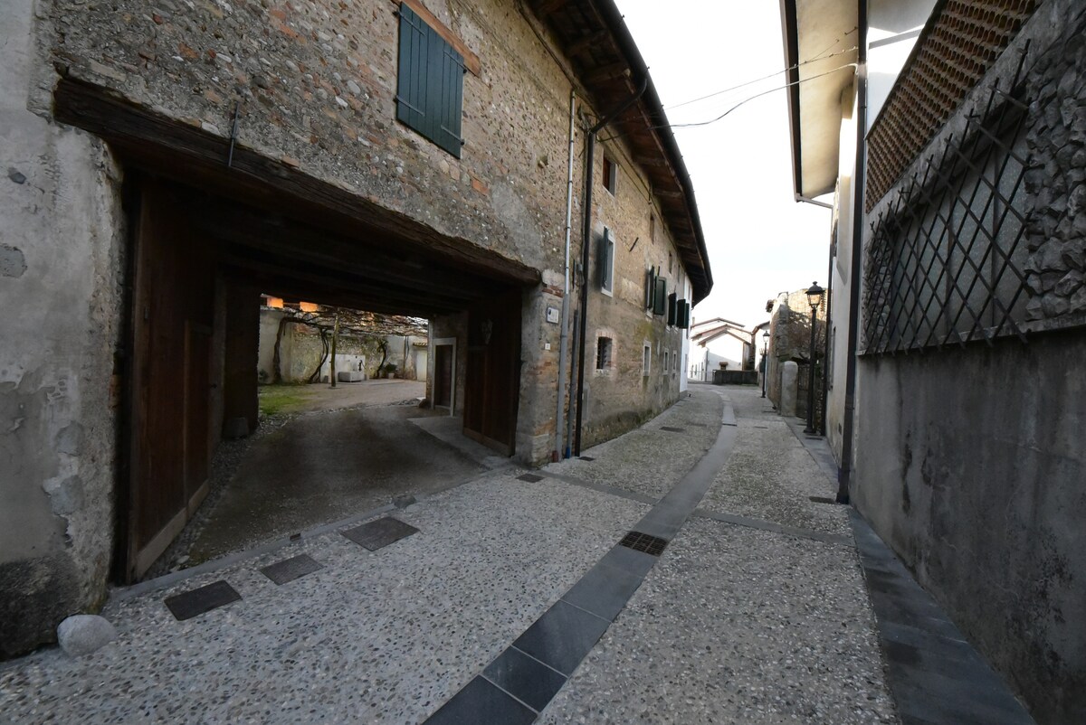 An inviting stone pathway runs alongside rustic buildings, characterized by textured walls and green window shutters. A spacious courtyard can be seen in the background, leading to an open area that creates a serene atmosphere. The scene is set in a quiet rural environment.