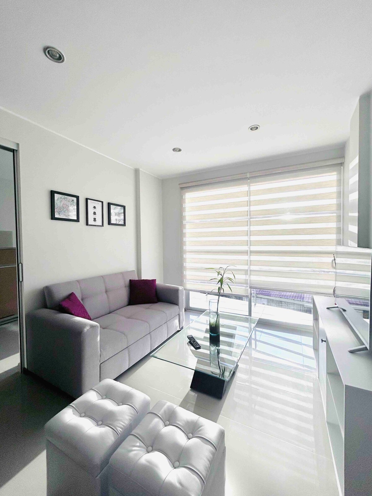 A modern living area features a gray sofa complemented by purple accent pillows. A glass coffee table sits in front of the sofa, reflecting light from the large windows with gently filtered shades, which create a bright and airy ambiance.