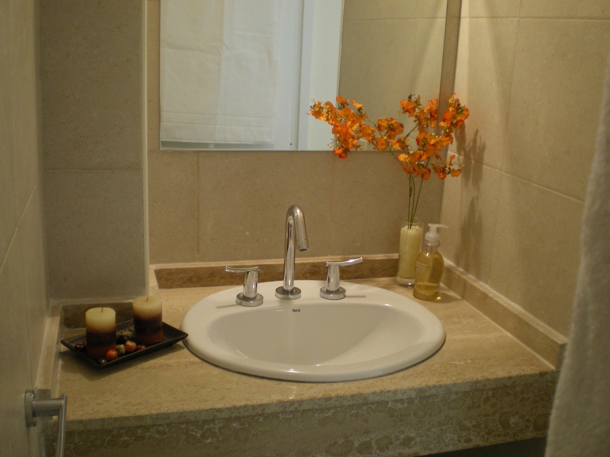 A bright bathroom sink area features a polished stone countertop with two candles and decorative flowers adding a touch of color. A soap dispenser and lotion bottle are positioned nearby, enhancing the practical and tidy appearance of the space.