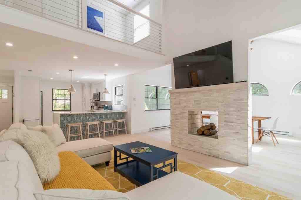 An open living area is featured, showcasing a light-colored sectional sofa and a blue coffee table. A sleek, modern fireplace is constructed from stone, with logs arranged inside. The kitchen area is visible in the background, highlighted by sleek bar seating and large windows allowing natural light.