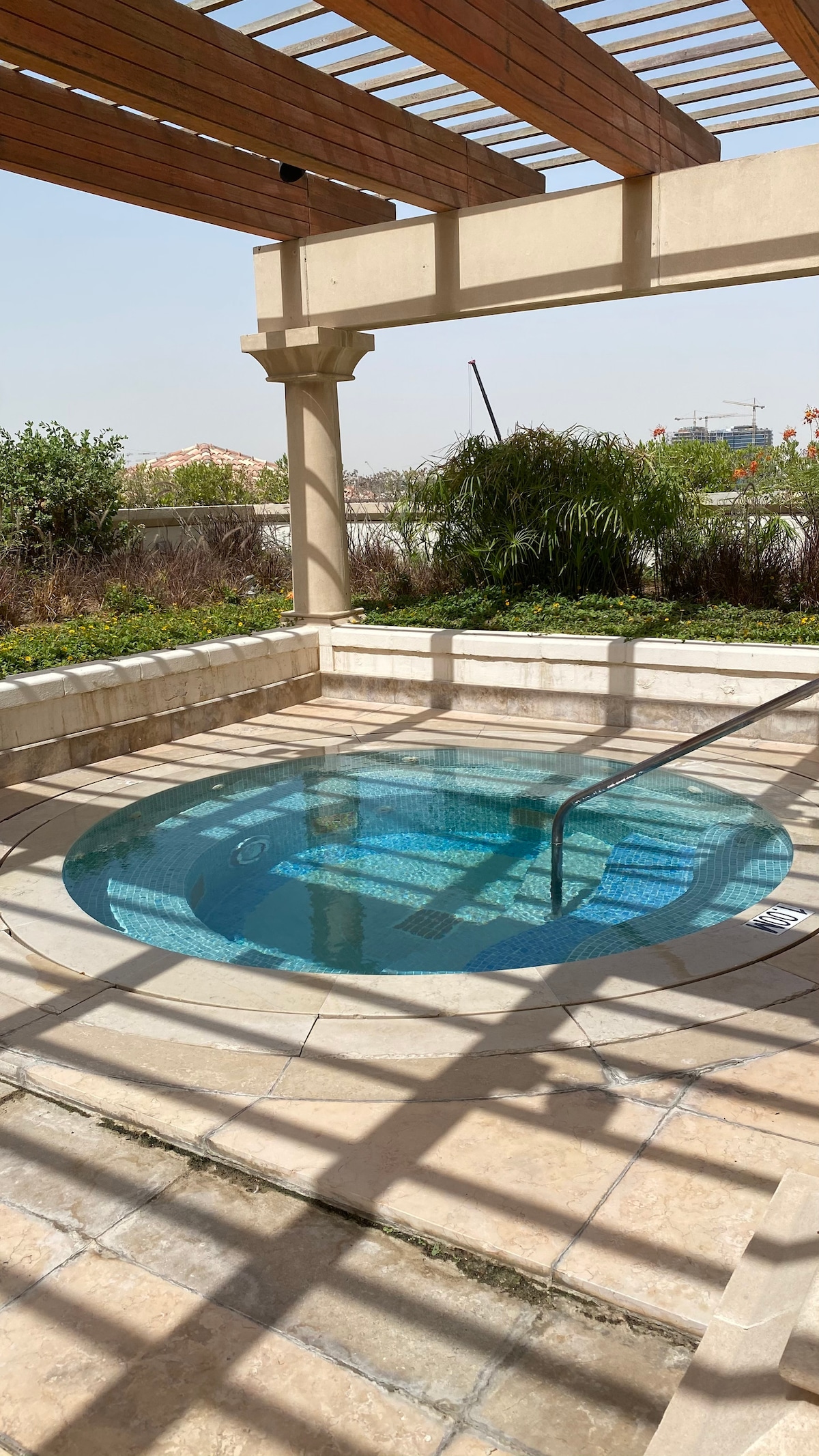 A private jacuzzi is surrounded by a tranquil outdoor space, with plants and greenery visible in the background. Natural light filters through a wooden trellis, casting intricate shadows onto the smooth stone terrace. The clear water of the jacuzzi reflects the serene environment.