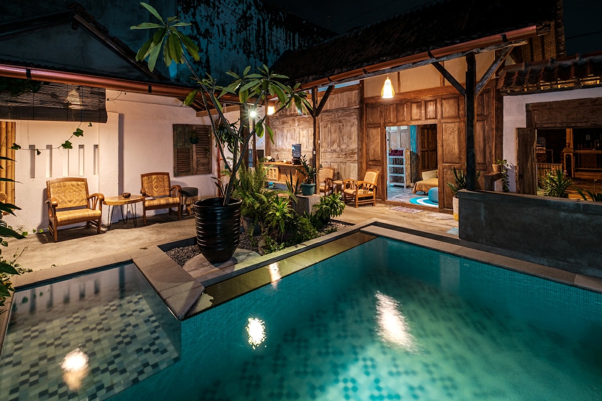 A tranquil outdoor space features a pool with clear water surrounded by tropical plants. Comfortable seating arrangements with wooden chairs are set near the entrance of the house, which exhibits traditional architecture with wooden doors. Soft lighting enhances the relaxed ambiance of the evening.