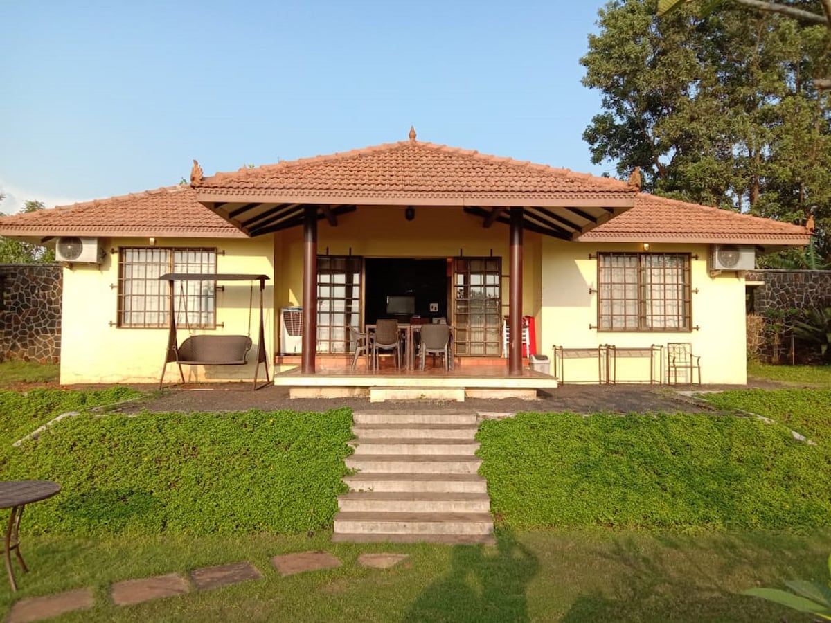 A charming villa is presented with a sloped roof and a wide front porch, featuring multiple seating arrangements. Large windows allow natural light to fill the interior. Steps lead from a grassy lawn to the entrance, adding to the welcoming appeal of the property.