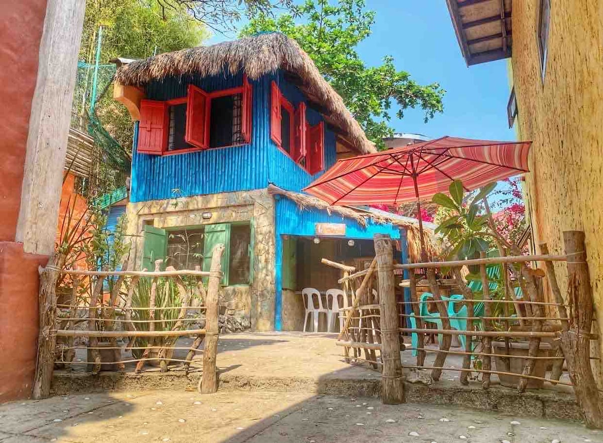 The vibrant two-story structure features a blue exterior with a thatched roof, accented by red-framed windows. A shaded outdoor area includes an umbrella and several white chairs, surrounded by lush greenery and rustic wooden fencing.