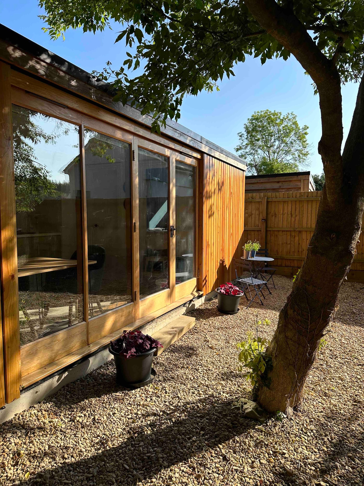 The cabin is surrounded by natural elements, featuring large glass sliding doors that allow ample light. A small outdoor seating area is present alongside a gravel pathway, with potted plants adding a touch of greenery. A tree provides shade, enhancing the peaceful atmosphere.