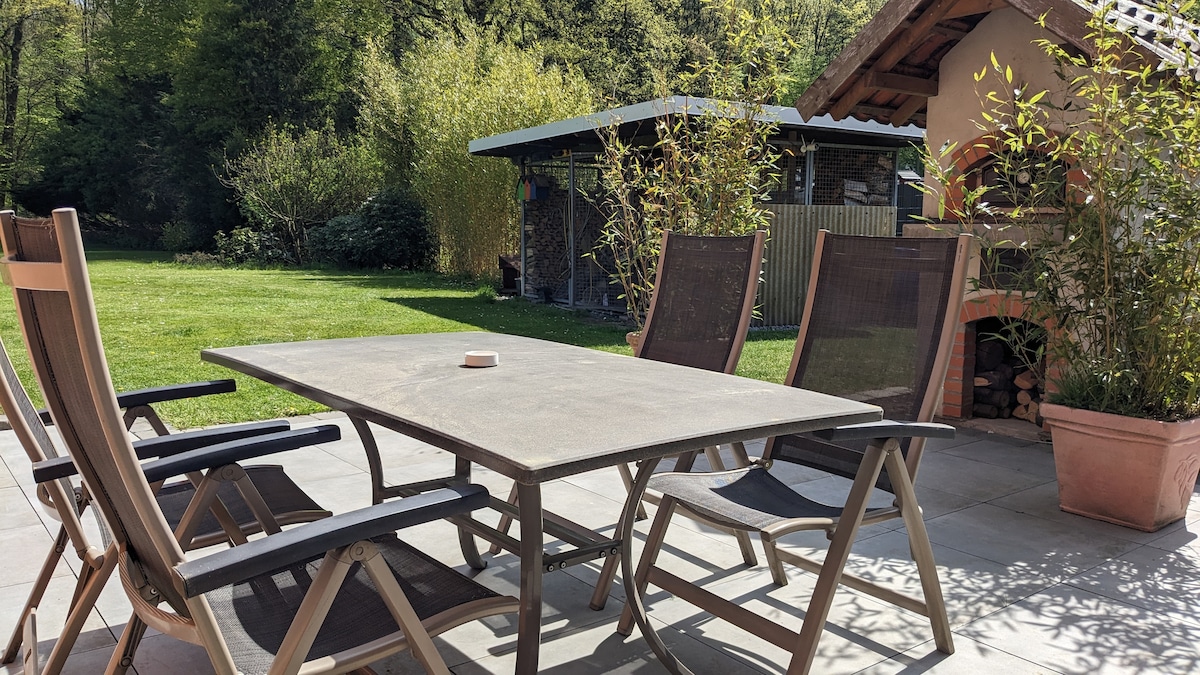 An outdoor terrace area is depicted, featuring a rectangular table surrounded by four wooden chairs with mesh backs. In the background, a well-maintained garden is visible, with a small shed nestled among greenery. The setting offers a peaceful view of the surrounding nature.