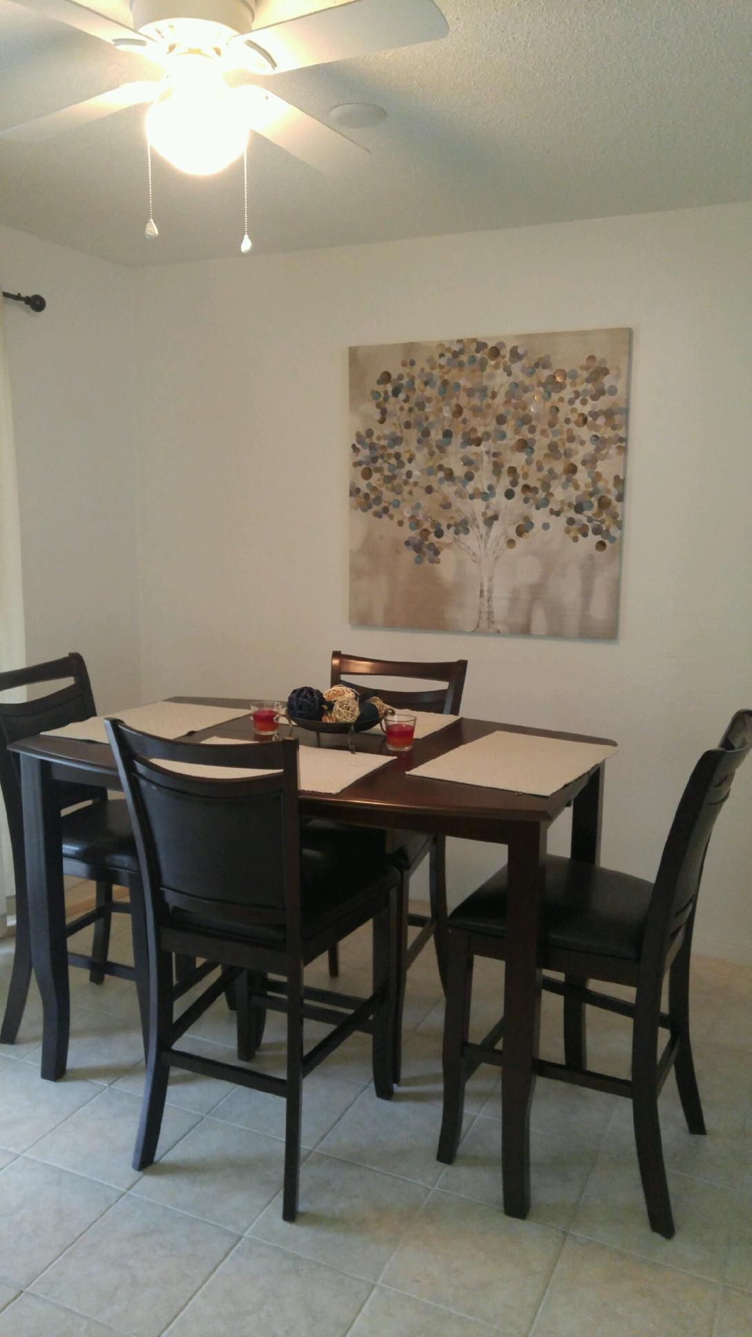 A dining area is depicted with a square table surrounded by high-backed chairs. A decorative piece showcasing a tree with gold and blue leaves adorns the wall. Soft lighting is provided by a ceiling fan, creating a calm and inviting dining space.