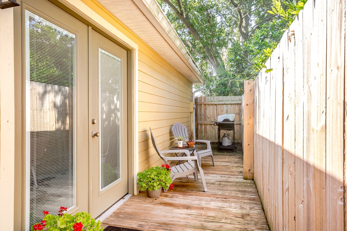 An outdoor wood deck is framed by a wooden fence, providing a private space. Two lounge chairs are positioned on the deck, accompanied by potted plants featuring vibrant flowers. Natural light filters through the surrounding trees, enhancing the serene atmosphere.