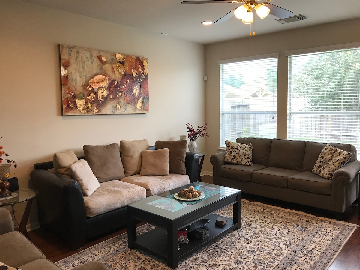 A cozy living room features two comfortable sofas with decorative pillows, arranged around a central coffee table. A large artwork with autumn leaves decorates one wall, while natural light enters through two windows with horizontal blinds. A patterned area rug adds warmth to the hardwood floor.