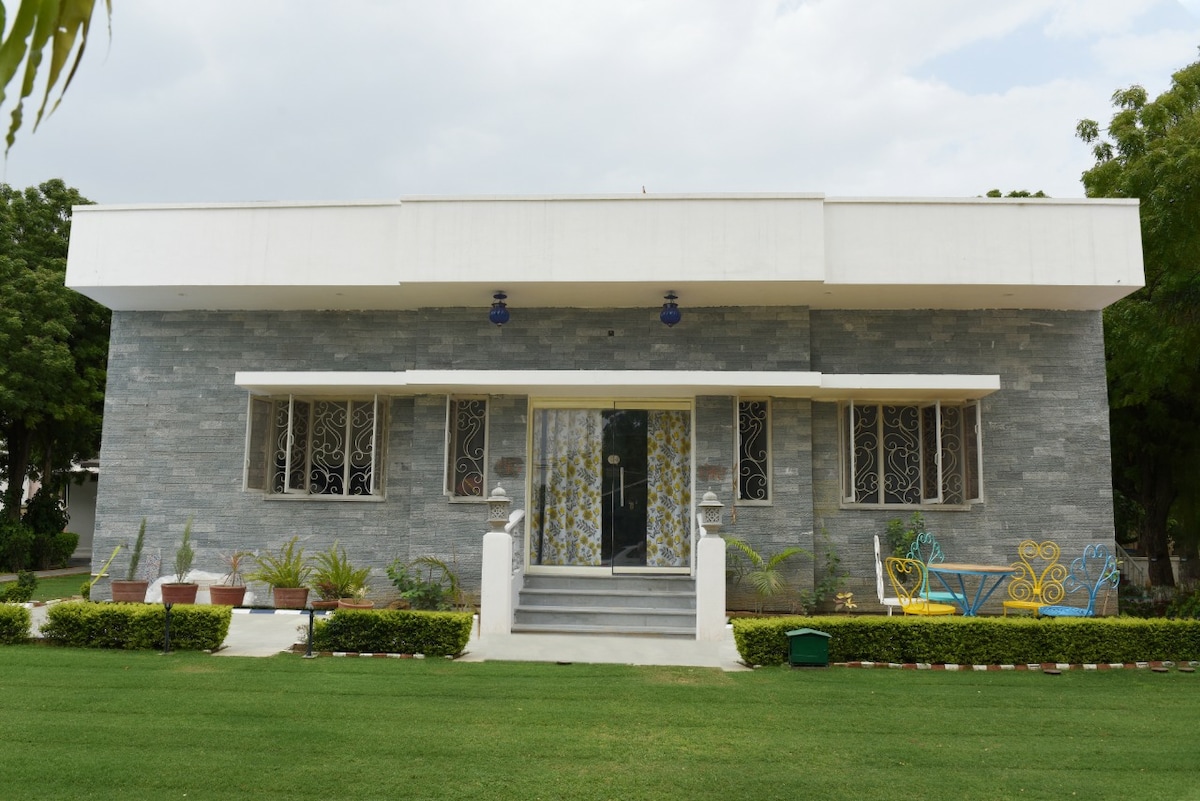 The exterior of the heritage home features a contemporary design with grey stone walls and large, decorative windows. A well-maintained lawn surrounds the building, complemented by potted plants. The entrance includes a staircase leading to a glass door, framed by a welcoming porch area.