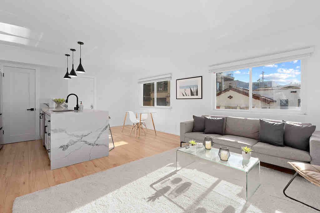 The modern kitchen features a spacious quartz island with seating and sleek stainless steel appliances. Large windows provide natural light, and a comfortable sofa is positioned near a glass coffee table. Neutral tones and simple décor enhance the inviting ambiance of the living area.