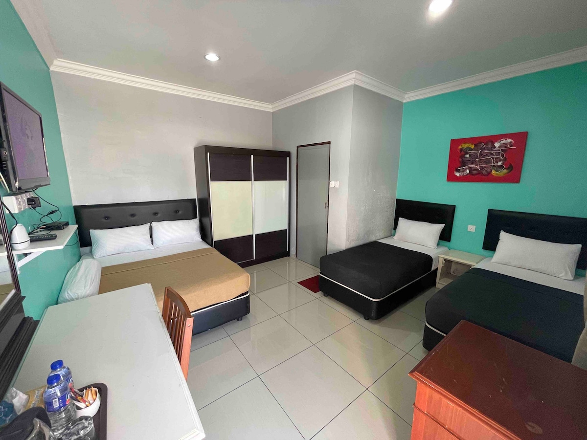 A room is presented with three beds arranged strategically, featuring a double bed, a single bed, and a stylish black bed. A modern wardrobe and a desk are included, while a television is mounted on the wall. Light walls are contrasted by vibrant turquoise accents.