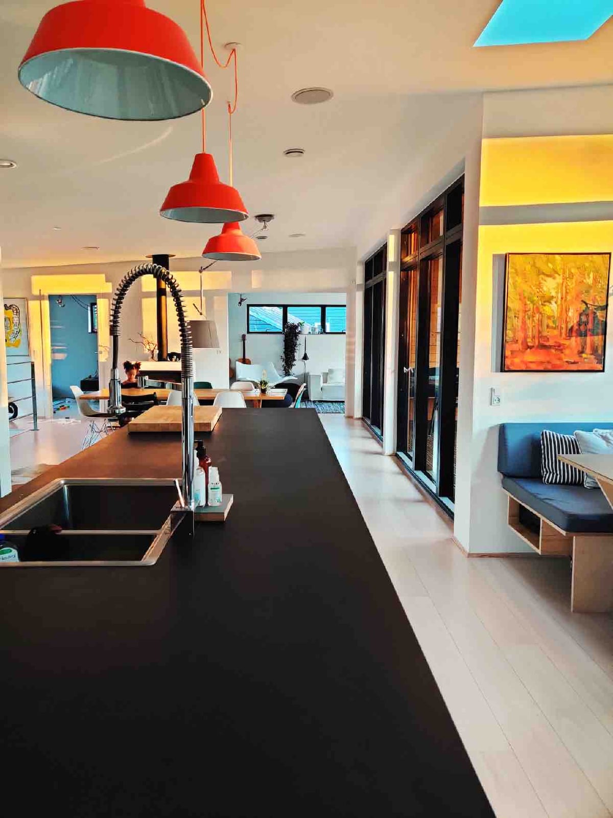 A modern kitchen area is visible, featuring a sleek black countertop complemented by stylish red pendant lights. Large glass doors provide a connection to the outdoor space, while a cozy seating area is seen in the background, illuminated by soft, warm lighting.
