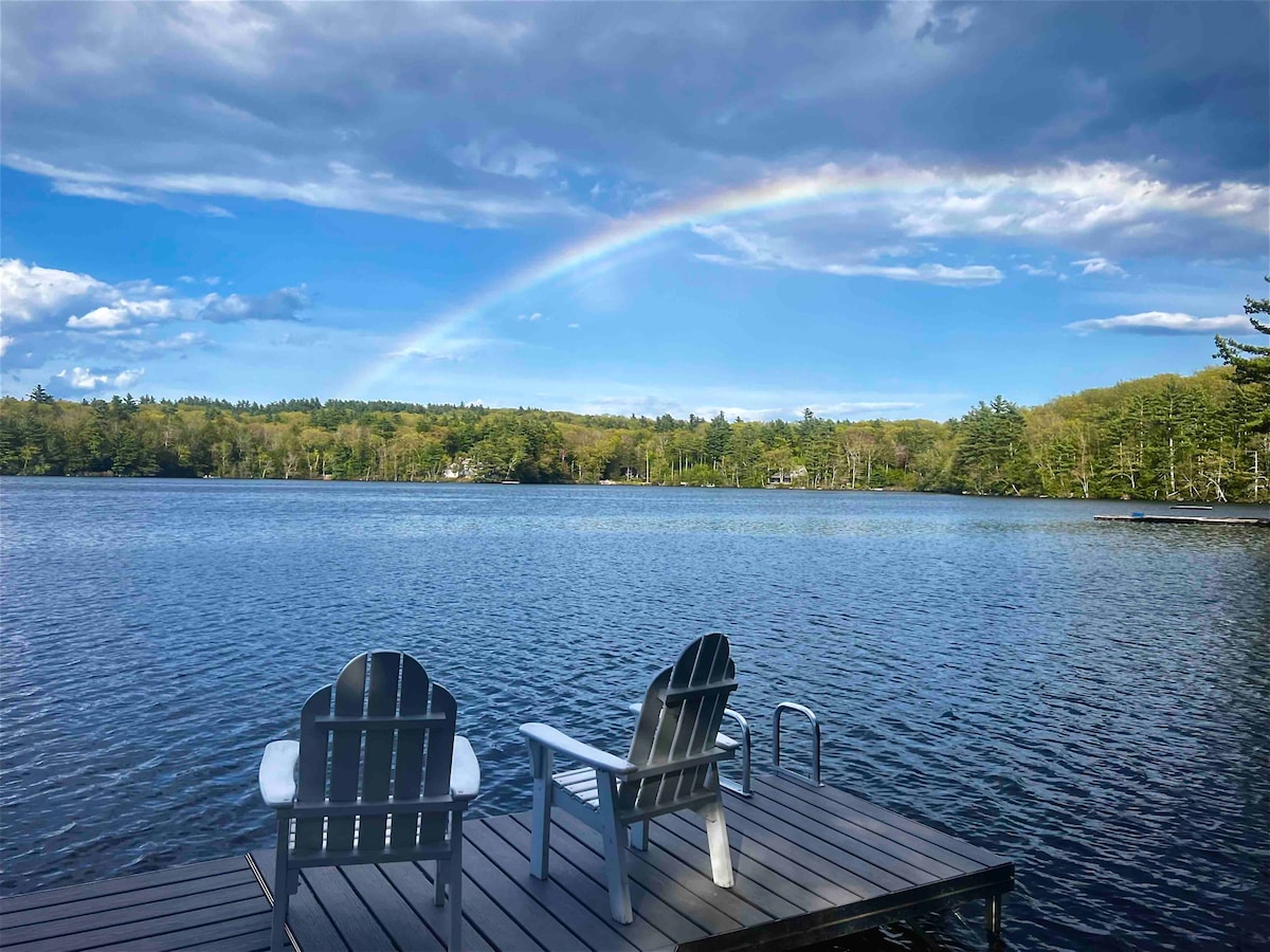 A wooden dock extends over the calm lake, featuring two Adirondack chairs positioned for lakeside relaxation. A vibrant rainbow arcs across a partly cloudy sky, while lush greenery frames the tranquil water, highlighting the natural beauty of the surroundings.