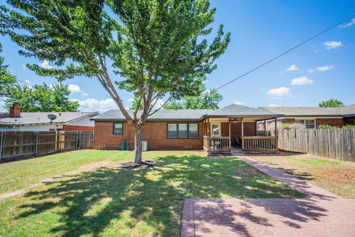 A well-maintained exterior features a single-story brick house surrounded by a spacious grassy yard. A large tree provides shade, while a paved walkway leads to a covered porch area at the entrance. The surrounding wooden fence offers privacy.