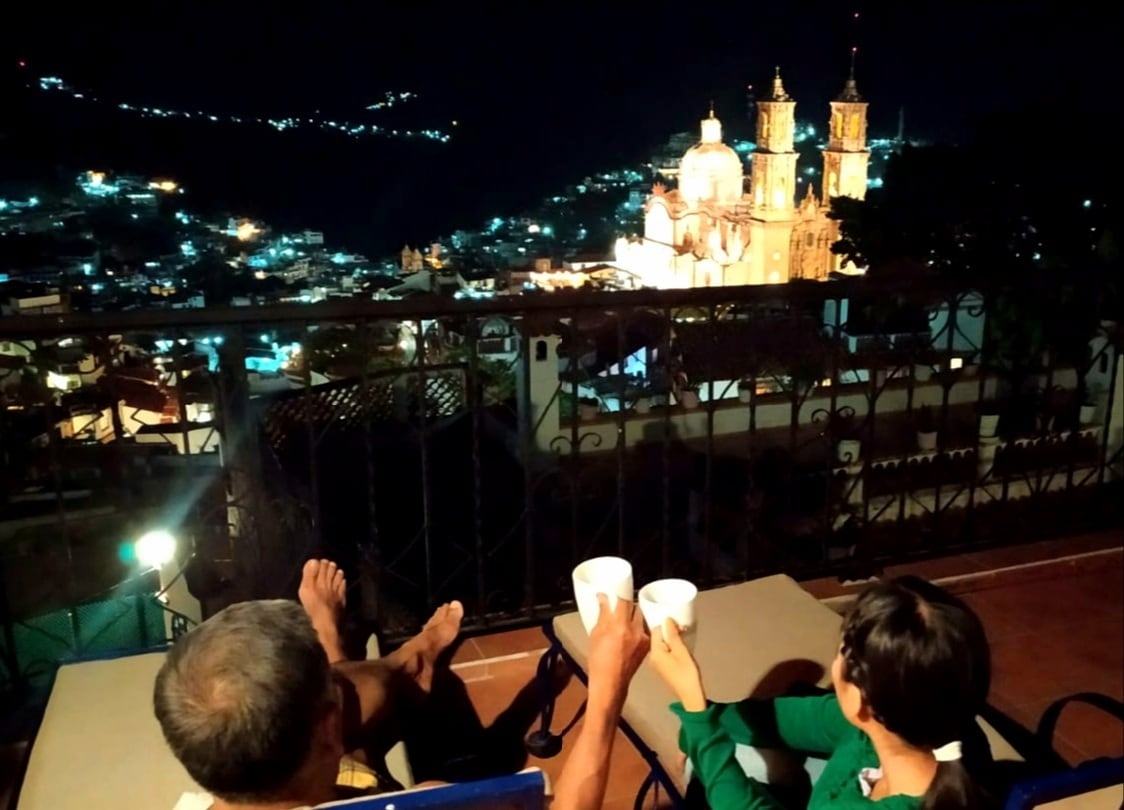 A nighttime view from the terrace showcases the illuminated Santa Prisca Church, with twinkling city lights in the background. Two guests are seated comfortably, holding cups, on outdoor chairs set on a wooden deck, enjoying the serene atmosphere.