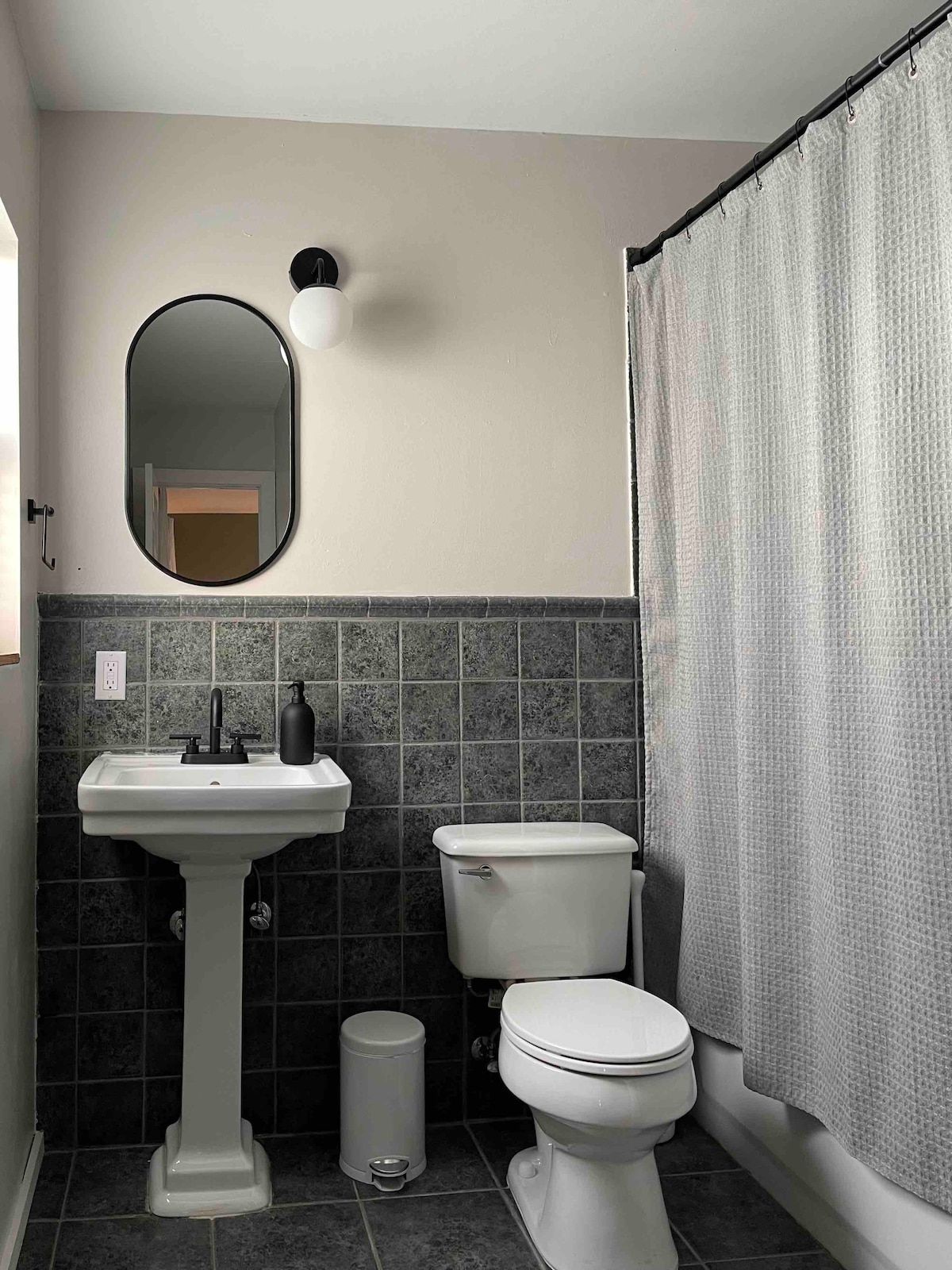 The bathroom features a white pedestal sink and a toilet, both positioned against a backdrop of dark tiled walls. A shower area is enclosed by a light grey curtain. A round mirror above the sink is complemented by a wall-mounted light fixture, enhancing the space's functionality.