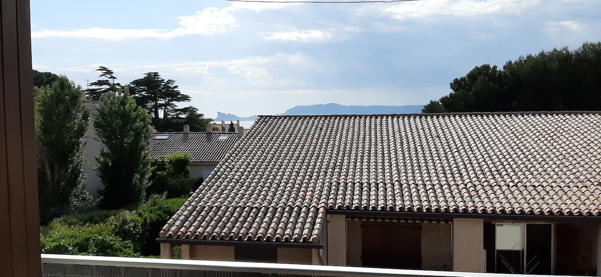 A view from the balcony offers a glimpse of the sea and distant mountains, with rooftops and greenery framing the scene. The sky is partly cloudy, allowing natural light to illuminate the area.