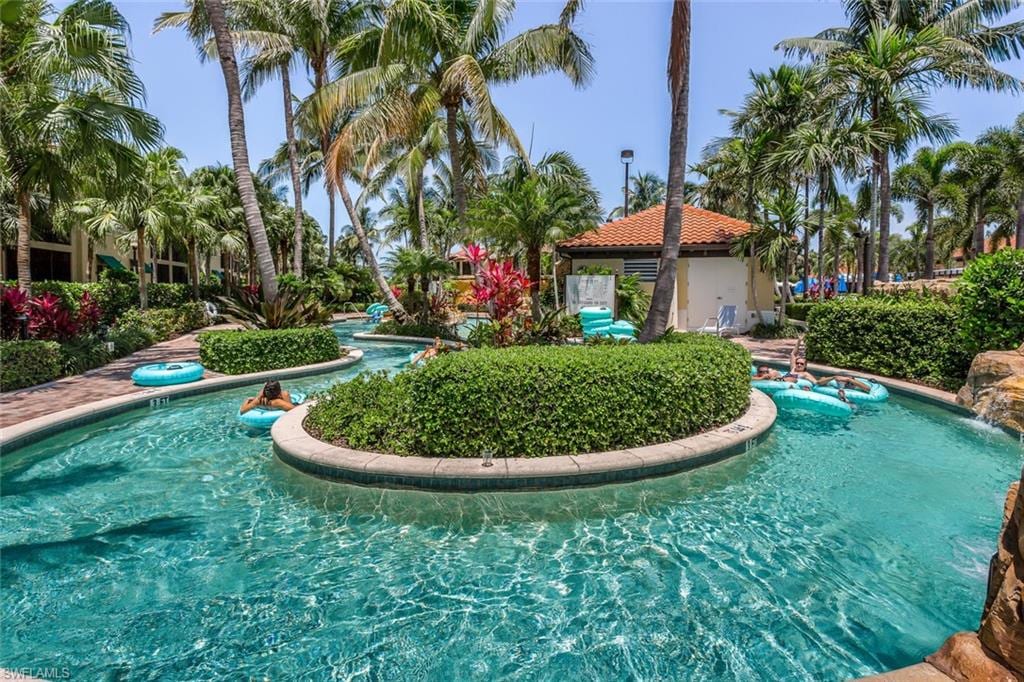 A winding lazy river pool is surrounded by lush palm trees and tropical landscaping. Colorful floats drift along the crystal-clear water, with vibrant flowers accentuating the serene atmosphere. A poolside building can be seen in the background, adding to the resort-like setting.