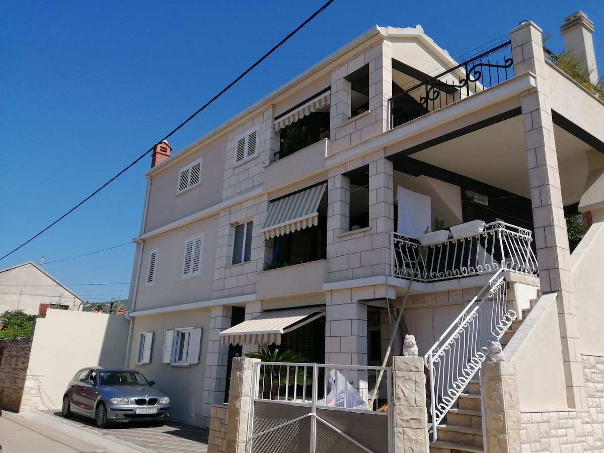 A contemporary building features a combination of stone and stucco façades. Spacious balconies are adorned with plants, and striped awnings provide shade. A staircase leads to a secured entrance, while a parked car is visible in the driveway, highlighting convenience for guests.