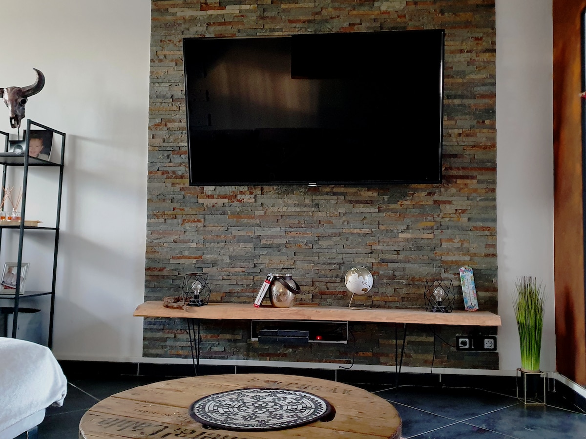 A modern living room features a mounted flat-screen television above a rustic wooden shelf, adorned with decorative items. A round coffee table with intricate designs sits on a dark tiled floor, contributing to the elegant ambiance of the space.